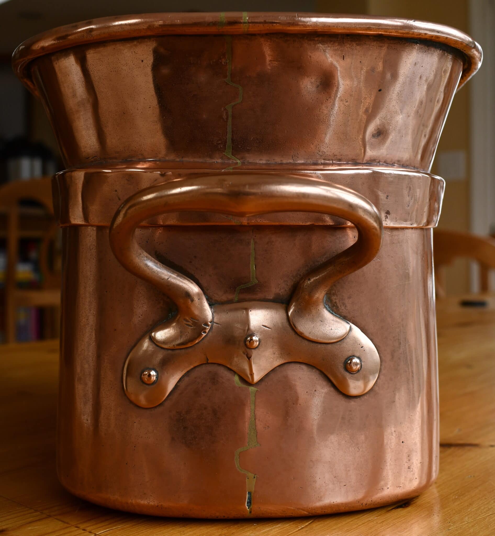 A tall copper stockpot (marmite) with two riveted handles, visible dents, and a vertical seam, on a wooden table.